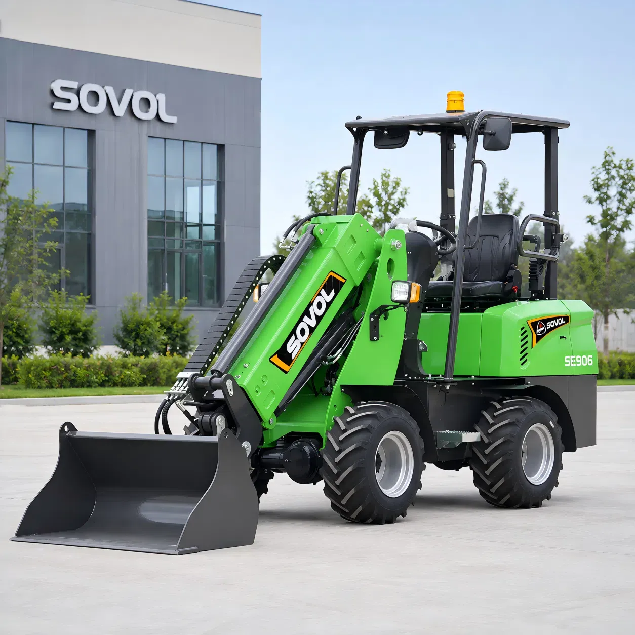 SOVOL SE906 elektrikli tekerlekli yükleyici, kepçe aparatıyla modern bir fabrika binasının önünde sergileniyor; çiftlikler, depolar, avlular ve hafif inşaat işleri için tasarlanmıştır.