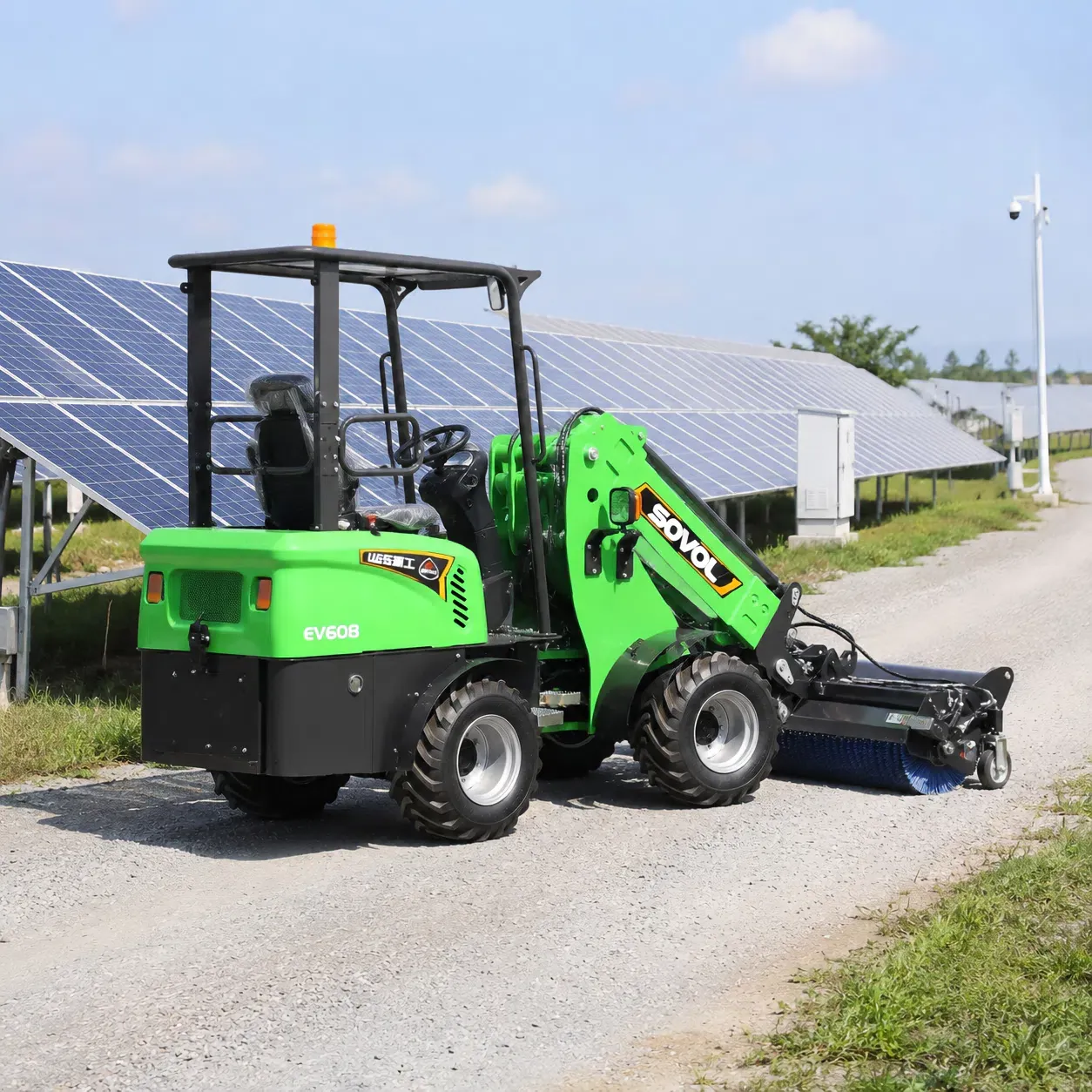 Vue latérale arrière de la chargeuse sur pneus électrique SOVOL SE906 équipée d'un accessoire de balayage, nettoyant un chemin d'accès en gravier entre les rangées de panneaux solaires dans un parc solaire.
